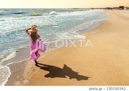Seascape on a sunny summer day. Woman on the beach, summer. Young happy woman carefree walking along the seashore in a pink fluttering dress, holding her hat on her head. View from above. 130180342