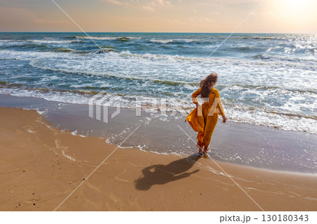 Seascape on a sunny day. Woman on the beach. Young woman in a yellow fluttering dress stands on the seashore. The girl looks at the sea Seascape on a sunny day. Woman on the beach. Young woman in a yellow fluttering dress stands on the seashore. The girl looks at the sea 130180343