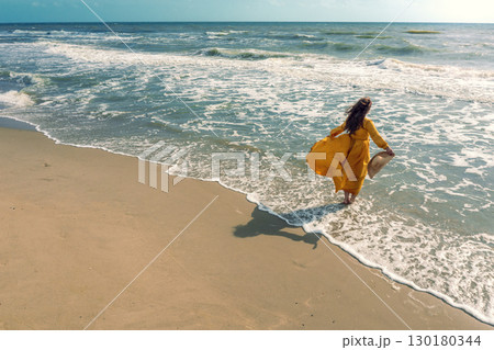 Seascape on a sunny day in summer. Woman on the beach, summertime. Young happy woman walks carefree on the seaside in yellow fluttering dress and sunhat. View from above 130180344