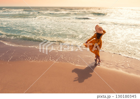 Seascape on a sunny day in summer. Woman on the beach, summertime. Young happy woman walks carefree on the seaside in yellow fluttering dress and sunhat. View from above Seascape on a sunny day in summer. Woman on the beach, summertime. Young happy woman walks carefree on the seaside in yellow fluttering dress and sunhat. View from above 130180345
