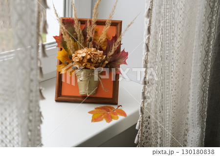 Handmade autumn decoration consisting of dried leaves and hydrangeas in small pot in wooden frame stands on windowsill. Autumn aesthetics 130180838