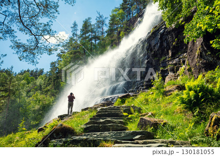 View from above of a beautiful waterfall cascade. Svandalsfossen (Sauda), Norway. Cascade creek of waterfall landscape on a sunny day. A man takes a photo of powerful waterfall 130181055