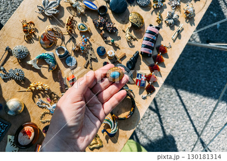 A hand selects a vintage brooch from a vibrant assortment of secondhand accessories laid out on a wooden table. Circular fashion, ethical adornment, reused beauty, zero-waste style 130181314