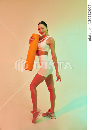 Orange colored yoga mat in hands. Young fitness woman in sportive clothes against background in studio Orange colored yoga mat in hands. Young fitness woman in sportive clothes against background in studio 130182007