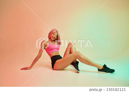 Sitting on the floor. Sportive woman with pink wig is against background 130182121