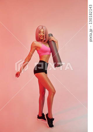 Holding yoga mat. Sportive woman with pink wig is against background Holding yoga mat. Sportive woman with pink wig is against background 130182143