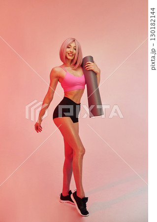 With yoga mat in hands. Sportive woman with pink wig is against background With yoga mat in hands. Sportive woman with pink wig is against background 130182144