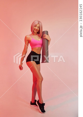 With yoga mat in hands. Sportive woman with pink wig is against background With yoga mat in hands. Sportive woman with pink wig is against background 130182145