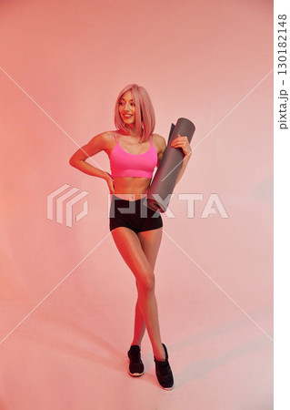 With yoga mat in hands. Sportive woman with pink wig is against background 130182148