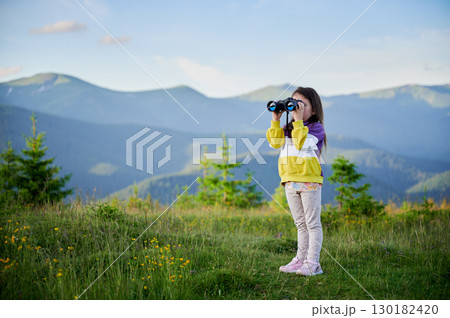 Young girl stands in grassy meadow, looking through binoculars, wearing yellow and purple sweatshirt. Behind her, rolling green hills and mountains stretch into distance under clear blue sky. 130182420