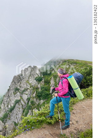 Woman hiker stands on misty mountain trail, gripping trekking poles. Female tourist with green sleeping pad strapped backpack and gazes at rugged, rocky cliffs partially shrouded in fog. 130182422