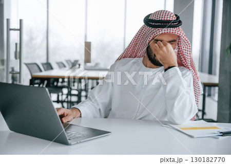 Feeling tired. Sitting by table with laptop. Successful Muslim businessman in traditional outfit in his office Feeling tired. Sitting by table with laptop. Successful Muslim businessman in traditional outfit in his office 130182770