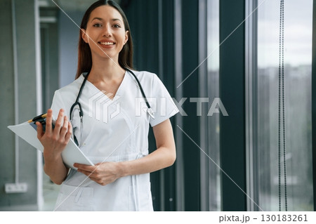Standing and holding documents in hands. Female doctor in white coat is indoors 130183261