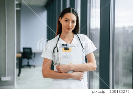 Holding notepad in hands. Female doctor in white coat is indoors Holding notepad in hands. Female doctor in white coat is indoors 130183273