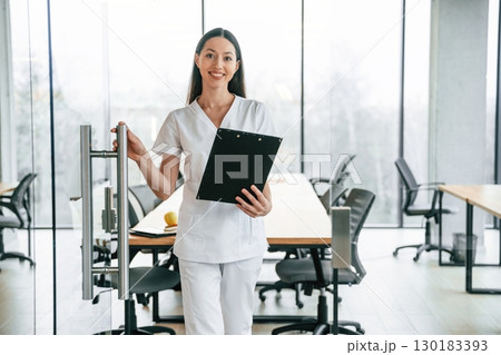 Front view. Standing with notepad in hands. Female doctor in white coat is indoors 130183393