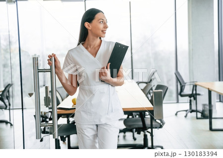 Front view. Standing with notepad in hands. Female doctor in white coat is indoors 130183394