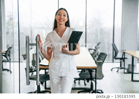 Front view. Standing with notepad in hands. Female doctor in white coat is indoors 130183395