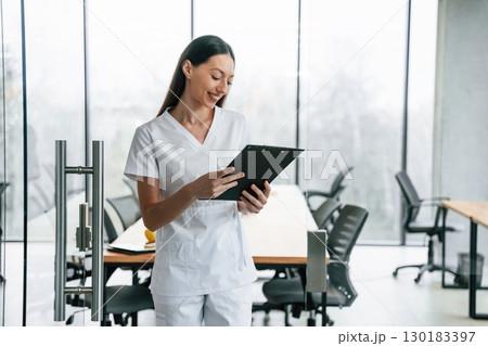 Front view. Standing with notepad in hands. Female doctor in white coat is indoors 130183397