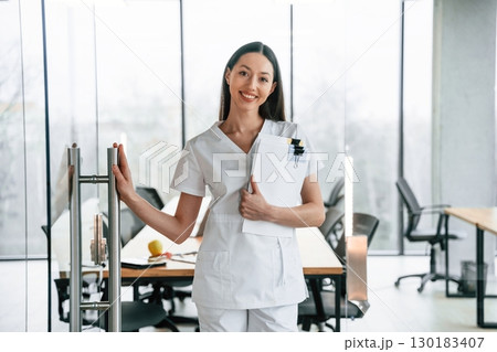 Leaving the meeting room. Standing with documents. Female doctor in white coat is indoors Leaving the meeting room. Standing with documents. Female doctor in white coat is indoors 130183407