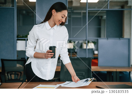 Holding drink in hand. Businesswoman is indoors in the office Holding drink in hand. Businesswoman is indoors in the office 130183535