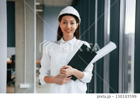 Plan and notepad in hands. Female architect in hard hat and in white clothes is indoors Plan and notepad in hands. Female architect in hard hat and in white clothes is indoors 130183599