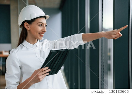Pointing by finger to the side. Female architect in hard hat and in white clothes is indoors Pointing by finger to the side. Female architect in hard hat and in white clothes is indoors 130183606