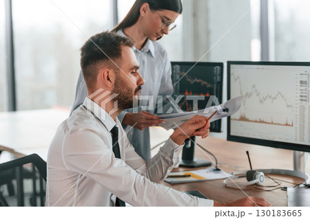 By the pc monitors. Two employees are working in the office together By the pc monitors. Two employees are working in the office together 130183665