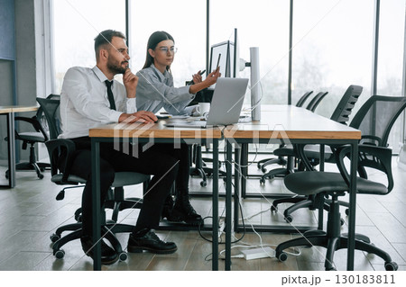 By the table with laptop. Two employees are working in the office together By the table with laptop. Two employees are working in the office together 130183811