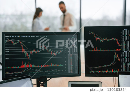 Standing behind the monitors with graphs on them. Two employees are working in the office together Standing behind the monitors with graphs on them. Two employees are working in the office together 130183834