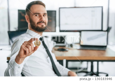 Showing golden coin that is in hand. Businessman is sitting by computer and working in the office Showing golden coin that is in hand. Businessman is sitting by computer and working in the office 130183946
