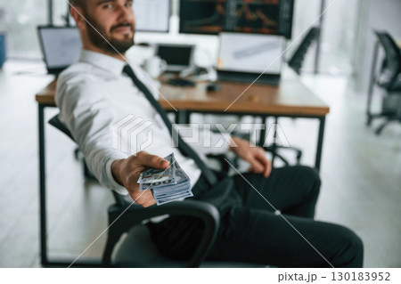 Giving lot of money. Holding in hand. Businessman is sitting by computer and working in the office 130183952