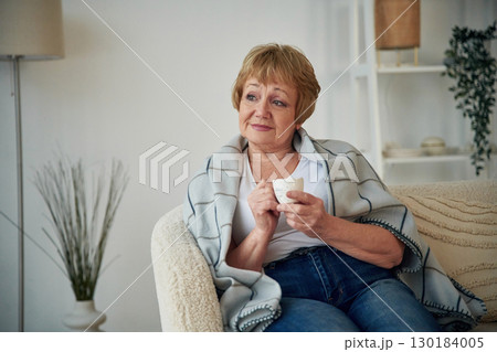 Cup with drink, sitting on a comfortable sofa. Senior elderly woman is at home in the living room Cup with drink, sitting on a comfortable sofa. Senior elderly woman is at home in the living room 130184005