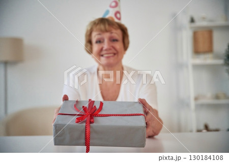 Smiling, celebrating birthday, holding gift box. Senior elderly woman is at home in the living room 130184108
