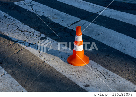 An orange traffic cone sits on cracked pavement at a busy crosswalk under bright sunlight in an urban setting 130186075