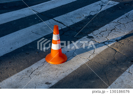 A solitary orange traffic cone is positioned on a damaged crosswalk marked by cracks in the surface, highlighting an urban setting 130186076