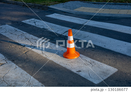 A bright orange traffic cone sits on a cracked crosswalk, marking a pedestrian area in a busy urban environment during dawn 130186077