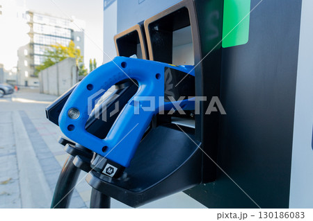 A blue fuel nozzle is positioned in a fuel pump at a contemporary gas station, surrounded by sleek buildings and sunlight 130186083