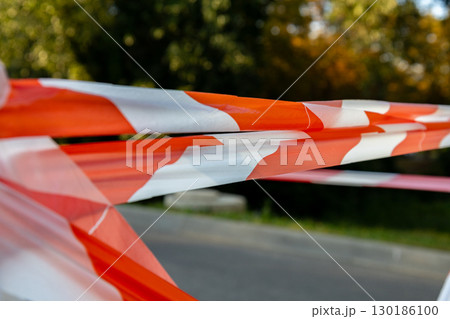 Bright orange and white barrier tape stretches across a path in a park, signaling a restricted area under vibrant trees during midday 130186100
