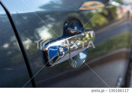 Close-up of a shiny car door handle reflecting sunlight showcases its sleek design and the vibrant surroundings of a suburban driveway 130186117