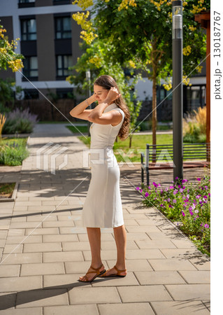 Young woman wearing a white midi dress walking outdoors in Old Money style. Concept of timeless fashion, graceful femininity and understated elegance in modern lifestyle. 130187767