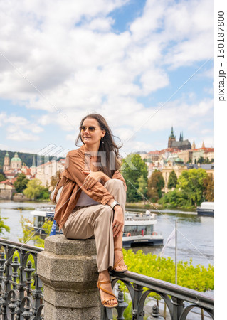 Young woman with a small backpack sitting on a stone railing with Prague Castle in the blurred background. Concept of solo travel, peaceful exploration and discovering historical European cities Young woman with a small backpack sitting on a stone railing with Prague Castle in the blurred background. Concept of solo travel, peaceful exploration and discovering historical European cities 130187800