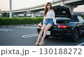 黒いオープンカーに腰かける若い女性｜夏の夕暮れの駐車場シーン 130188272