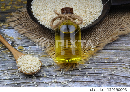 Glass bottle of sesame oil and raw sesame seeds in wooden spoon or bowl on rustic table. Uncooked sesame background concept with copy space Glass bottle of sesame oil and raw sesame seeds in wooden spoon or bowl on rustic table. Uncooked sesame background concept with copy space 130190008
