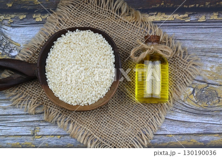 Glass bottle of sesame oil and raw sesame seeds in wooden spoon or bowl on rustic table. Uncooked sesame background concept with copy space 130190036