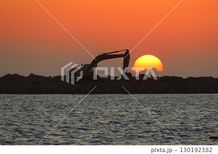An excavator silhouetted on rocks for marina construction at sunset. With the sun shining brightly. An excavator silhouetted on rocks for marina construction at sunset. With the sun shining brightly. 130192382