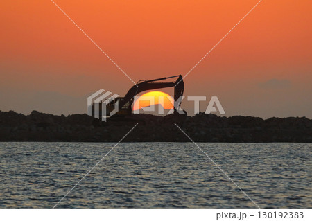 An excavator silhouetted on rocks for marina construction at sunset. With the sun shining brightly. An excavator silhouetted on rocks for marina construction at sunset. With the sun shining brightly. 130192383