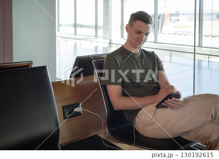 Young man charging phone at airport, using smartphone while waiting for flight. Comfortable travel concept in modern terminal. 130192615