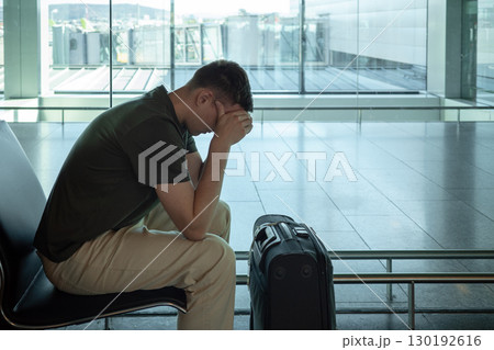 Young man with suitcase sitting in airport terminal looking stressed and disappointed. Concept of flight delay, cancellation or travel problems. 130192616