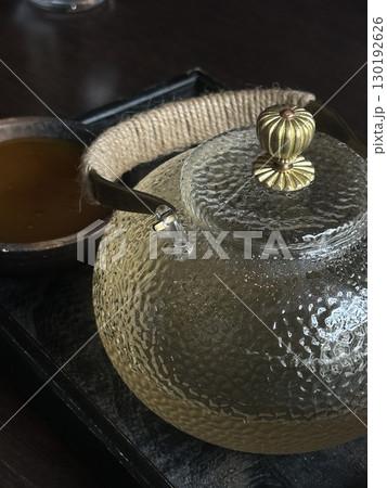 Close up of textured glass teapot with golden lid and rope handle on a wooden tray. Concept of tea ceremony, warmth, and cozy atmosphere. Close up of textured glass teapot with golden lid and rope handle on a wooden tray. Concept of tea ceremony, warmth, and cozy atmosphere. 130192626