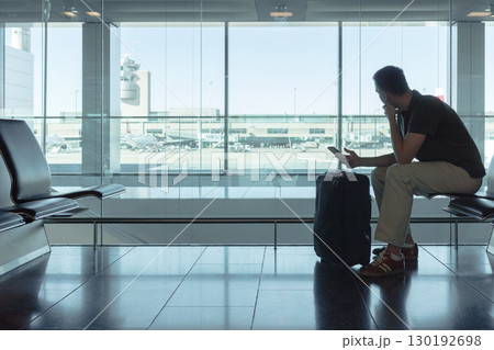 Young man sitting at airport gate waiting for a flight, looking out the window at airplanes with smartphone and suitcase. Modern air travel concept. Young man sitting at airport gate waiting for a flight, looking out the window at airplanes with smartphone and suitcase. Modern air travel concept. 130192698